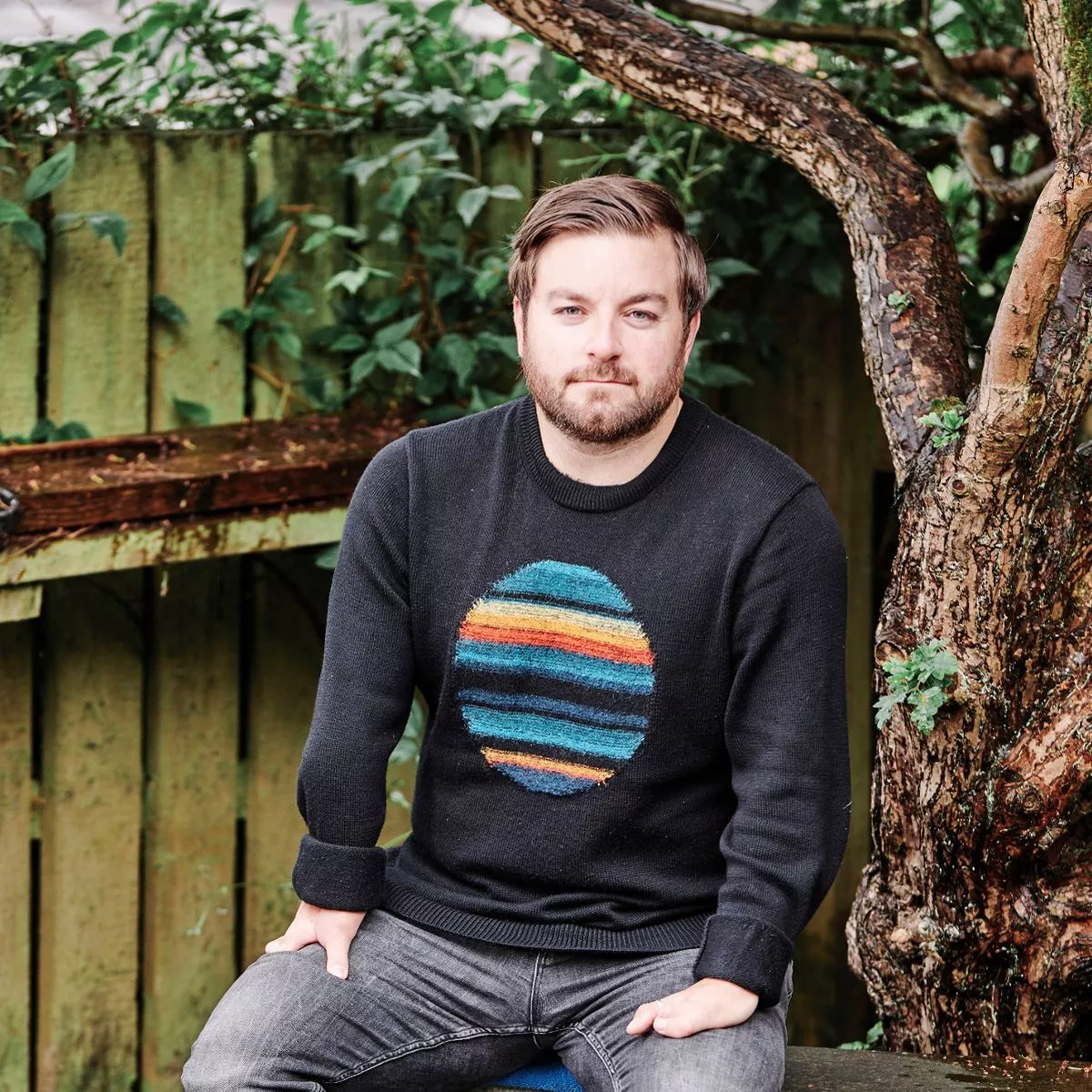 A photo of Alex Brooker sat in a tree. He is a white man with dark hair and beard and he is wearing a navy jumper