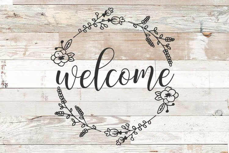 "Welcome" written in black with black flowers around the word. The background is rustic looking beach wood.