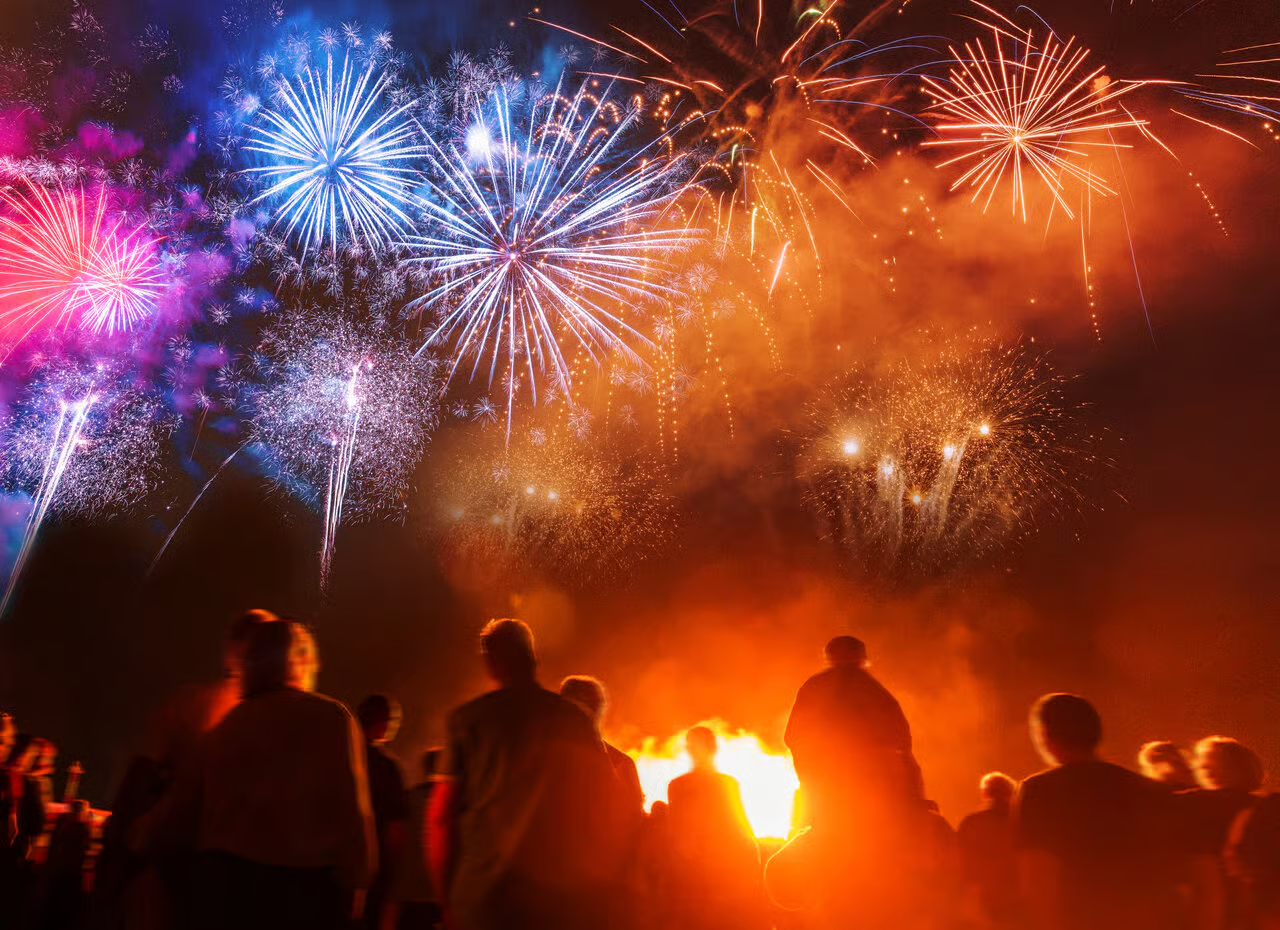 A community of people standing around a bonfire and watching an array of colourful fireworks.
