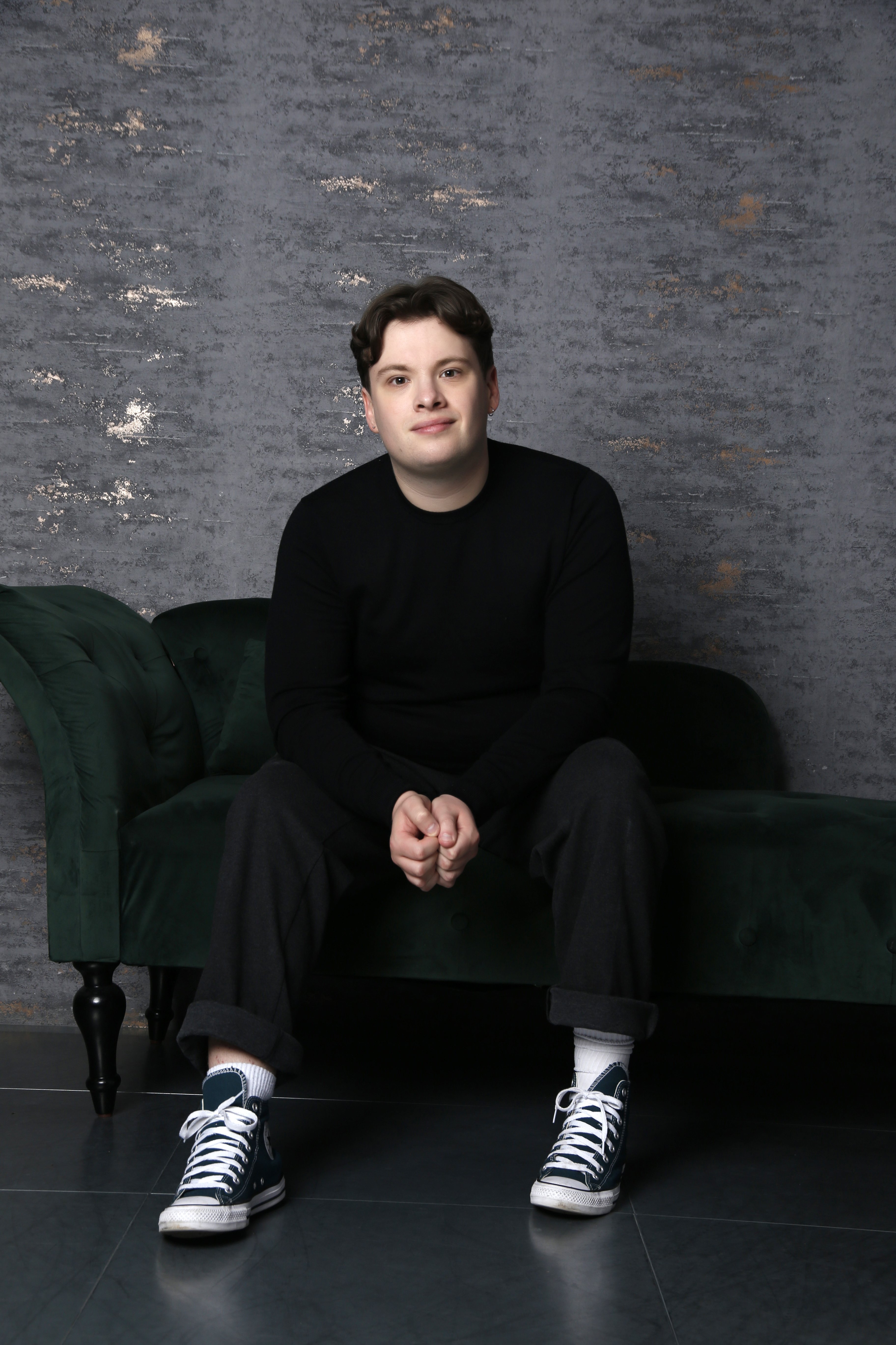 A man with short brown hair sat on a green sofa. He is wearing a black jumper and black trousers. Facing the camera.