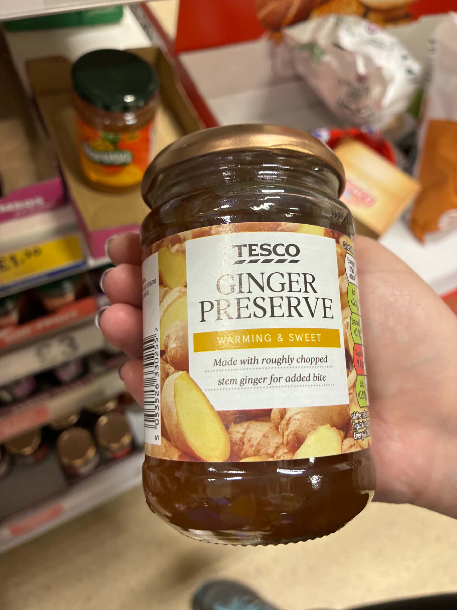 A glass jar of Tesco Ginger Preserve held in a person's hand. The label reads "TESCO GINGER PRESERVE", described as "WARMING & SWEET", and notes "Made with roughly chopped stem ginger for added bite" against a background image of sliced ginger root