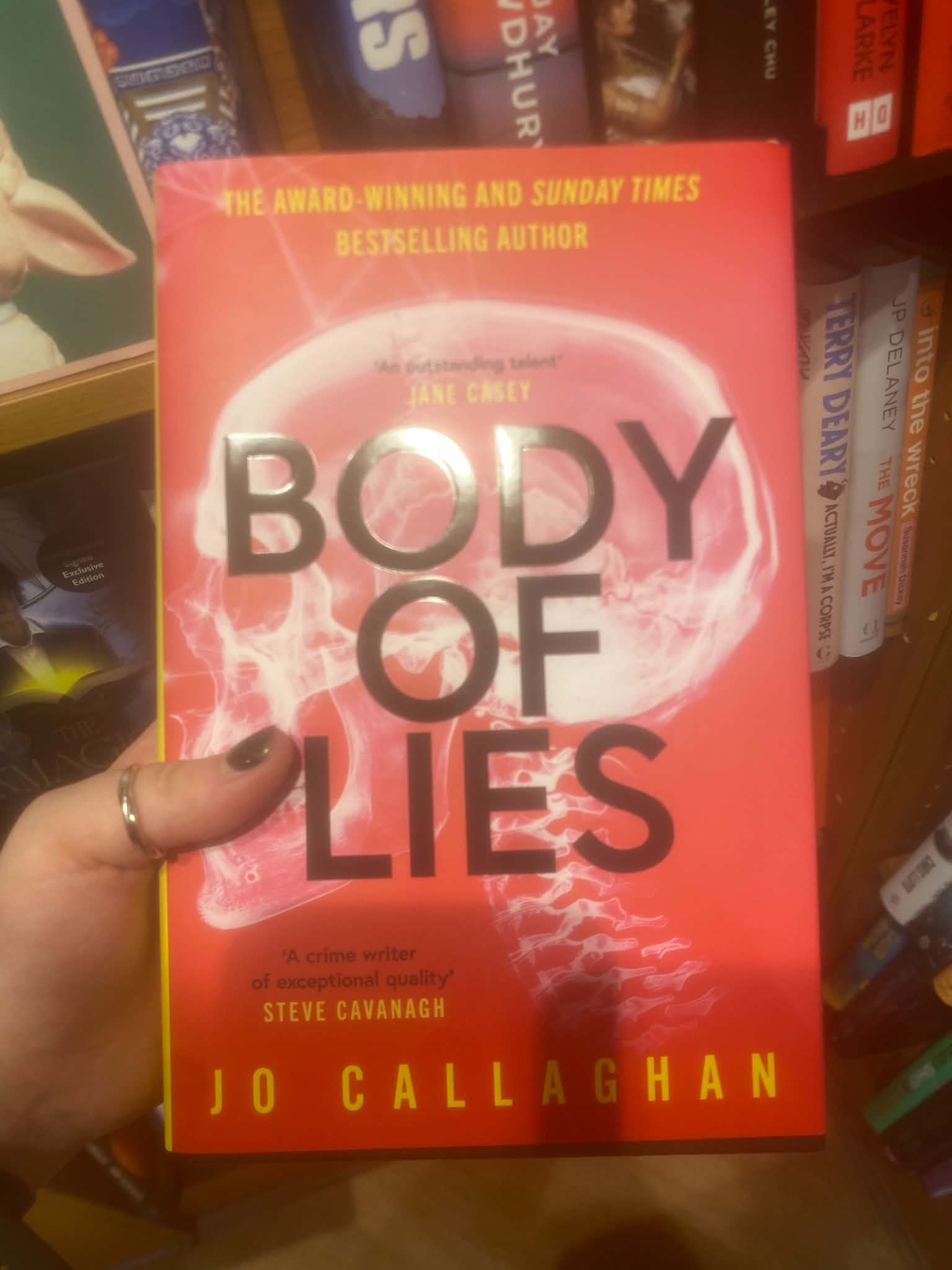 A hand holding the red paperback cover of the novel Body of Lies by Jo Callaghan.