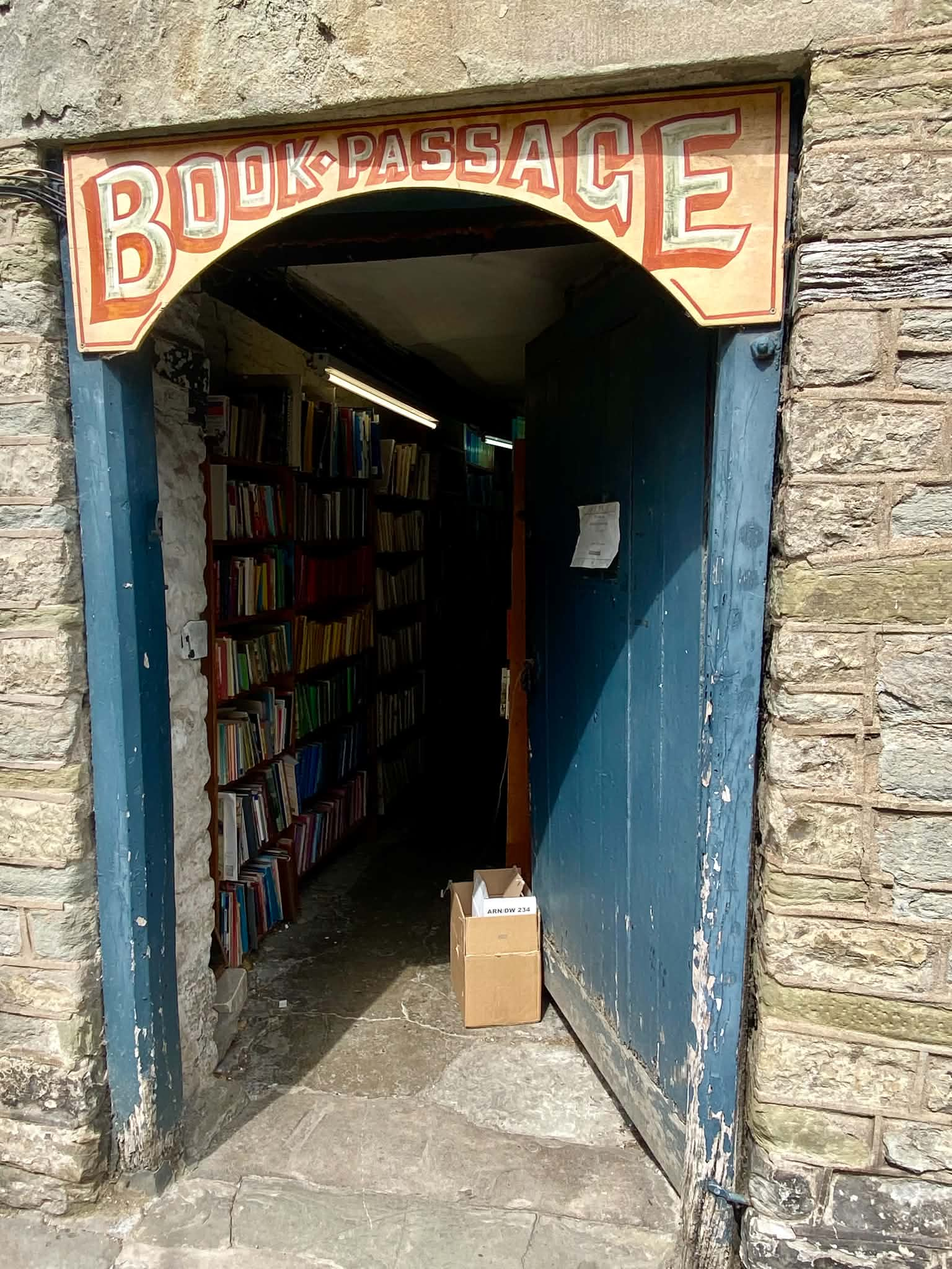 The stone arch entrance of a narrow shop with an arched sign reading "BOOKPASSAGE" overhead, leading into a passageway lined with fully packed bookshelves.