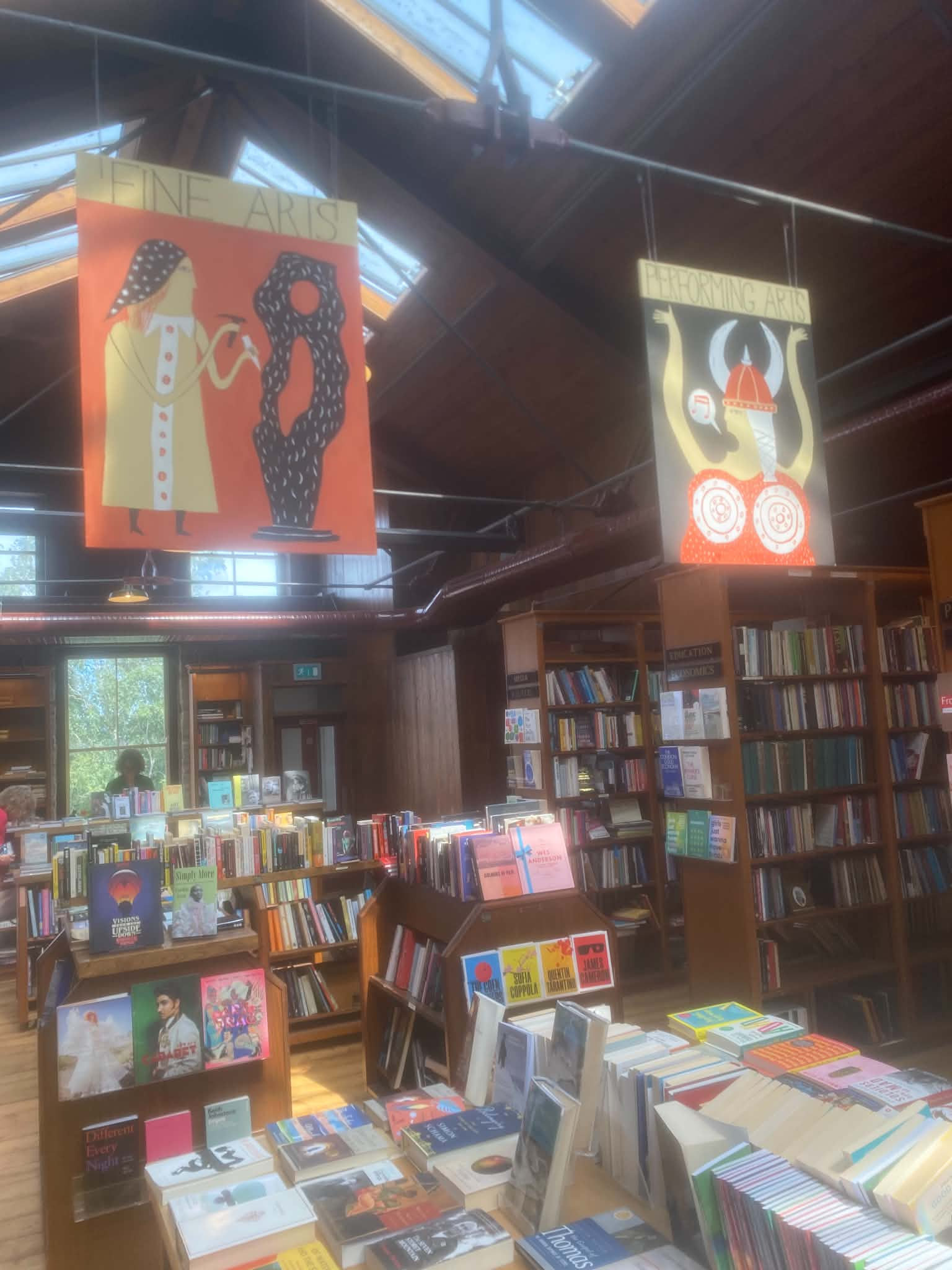 iThe interior of a spacious bookstore with wooden ceilings, featuring central display tables loaded with books, tall surrounding bookshelves, and decorative banners hanging from above.