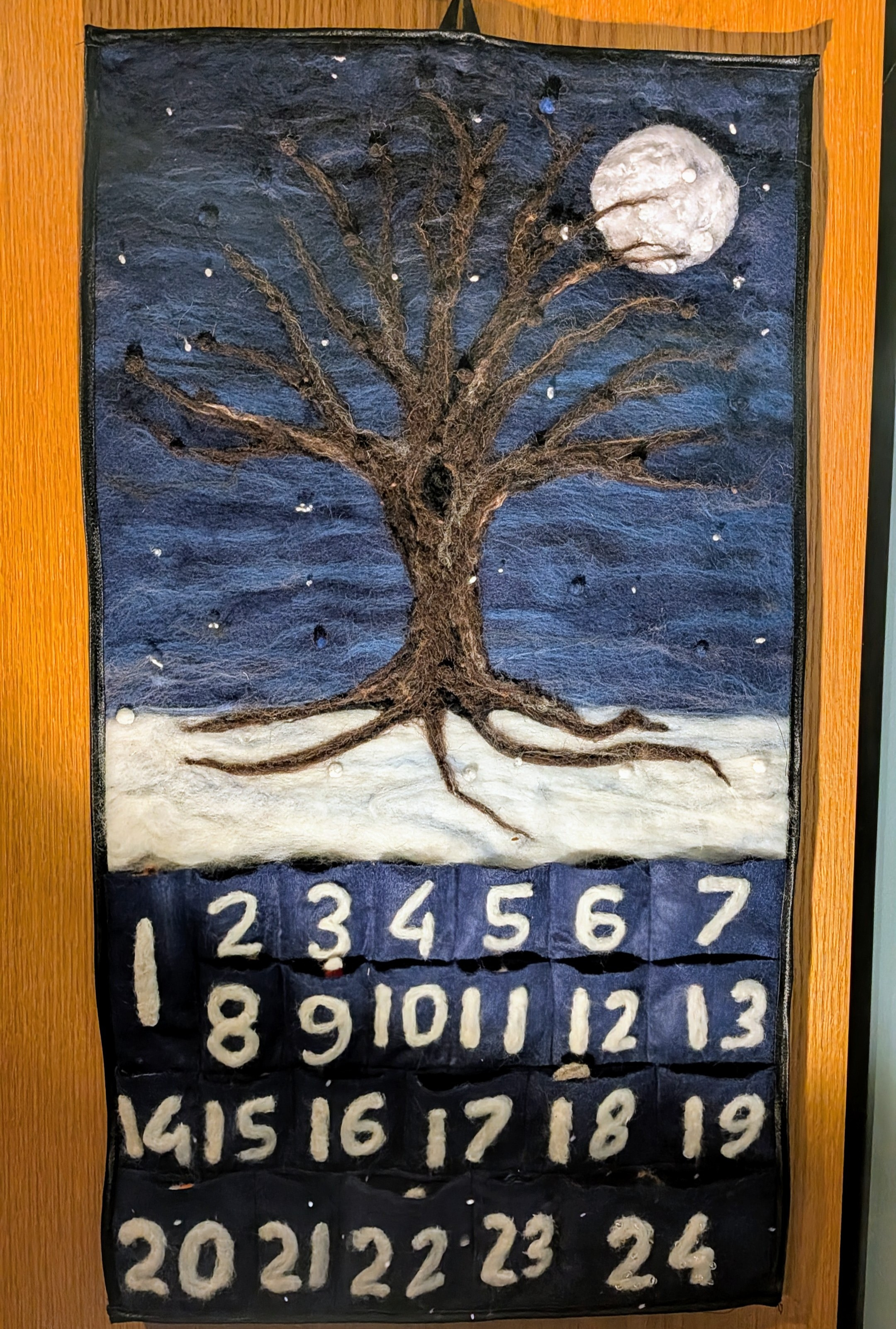 A needle felted winter tree advent calendar on a blue hazy background with snow underneath and the numbers 1 to 24