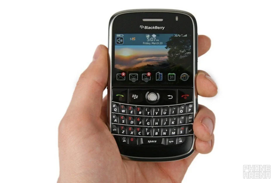 A hand holding a black BlackBerry smartphone with a full physical QWERTY keyboard, a navigation trackball, and a color screen displaying a sunset landscape wallpaper along with app icons.