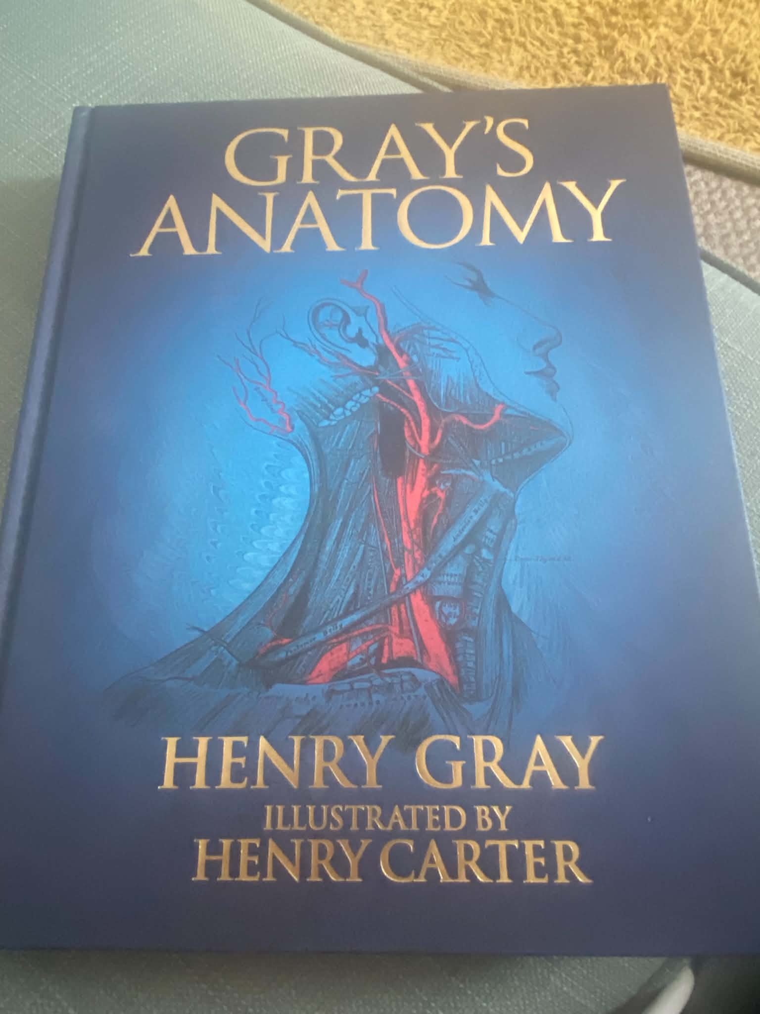 A close-up of a dark hardcover book titled "Gray's Anatomy" by Henry Gray, illustrated by Henry Carter, displaying a blue line-art anatomical illustration.