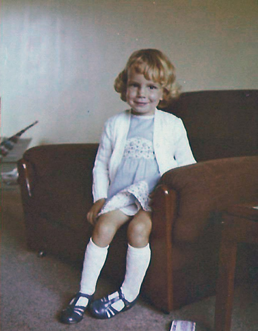 Young Sara age 4 sat on a brown armchair. She has short blond hair and is wearing a blue dress and white cardigan.