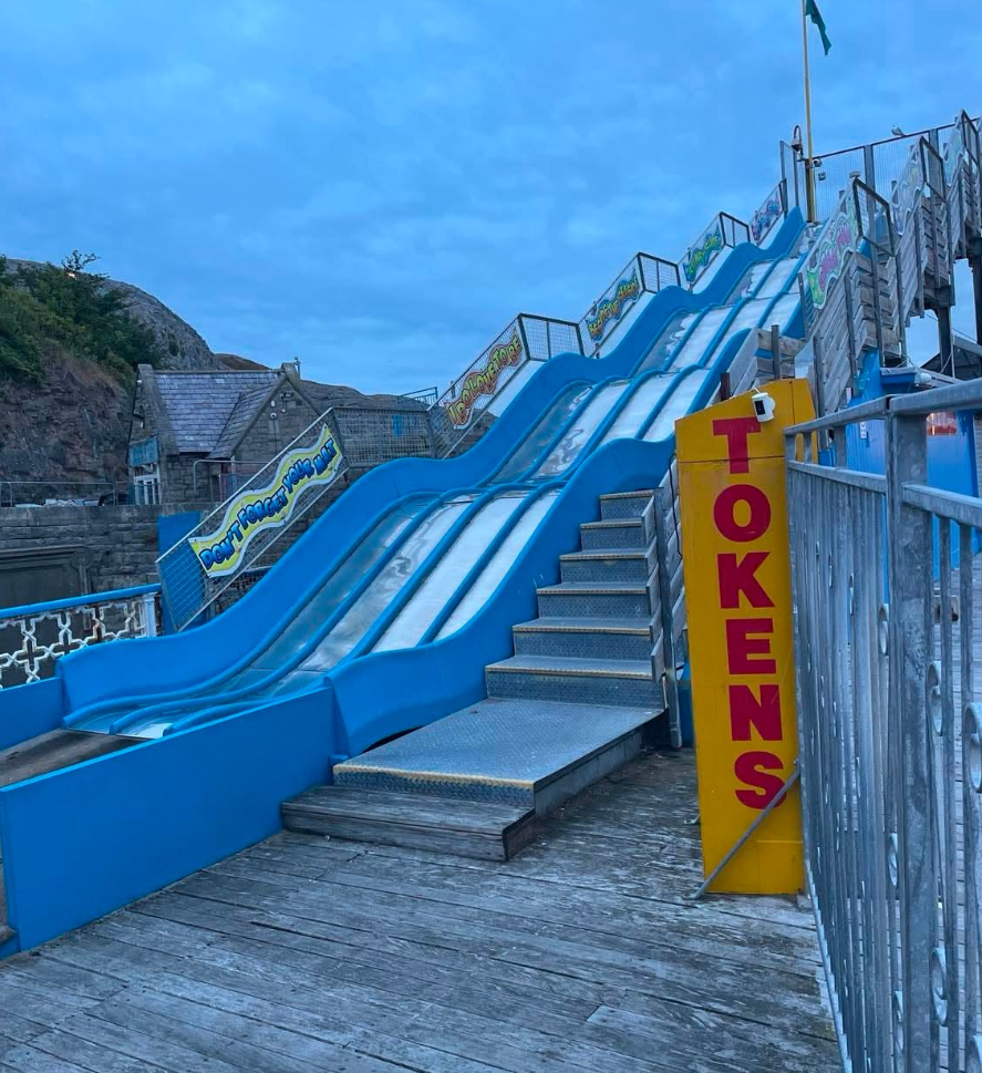 A large, multi-lane blue and silver helter-skelter slide on a wooden pier next to a tall yellow sign reading "TOKENS" in red lettering.