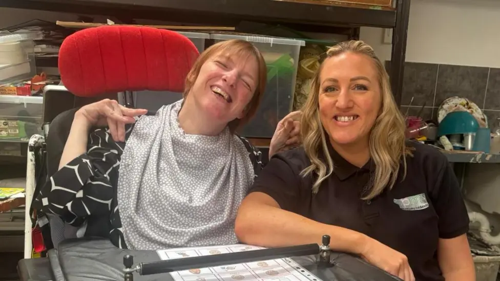 Sarah sit next to Vicky. Vicky is in her wheelchair. They are both smiling to the camera.