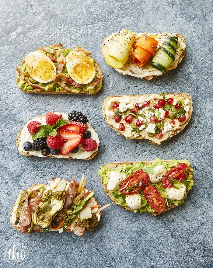 An overhead shot showcasing a variety of colourful, gourmet toast combinations arranged in two columns on a grey surface. The toppings feature a mix of fresh fruits, berries, vegetables, eggs, meats, and cheeses on toasted sliced bread.