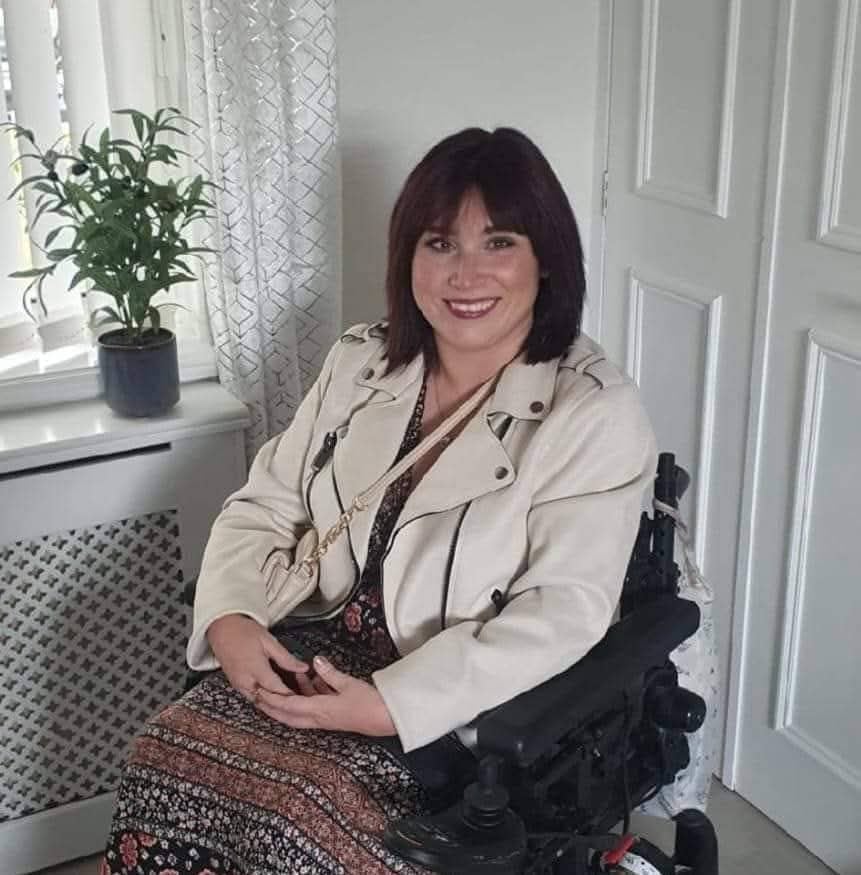 A white female with shoulder length brown hair. She is wearing a short cream jacket and brown and black long skirt. She is sitting in a wheelchair.
