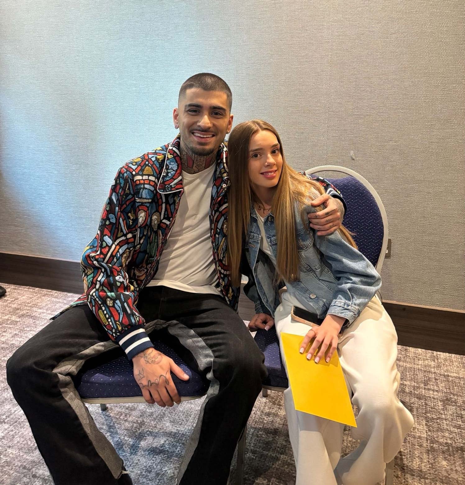 A young black man wearing a rainbow style jacket, sat next to a teenage girl with long blond hair. She is wearing a denim jacket and white trousers.
