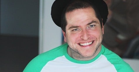 Andrew is smiling to the camera. He is wearing a black hat. He has a green and white top.