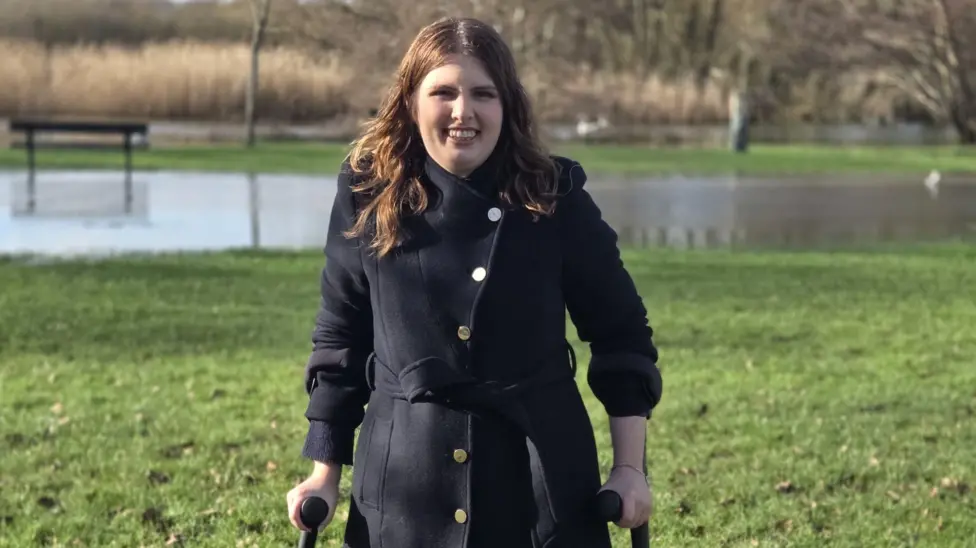 A person standing outdoors on a grassy area, using two crutches. They are wearing a dark coat with gold buttons. Behind them is a large puddle of water, a bench, and trees on a cloudy day.
