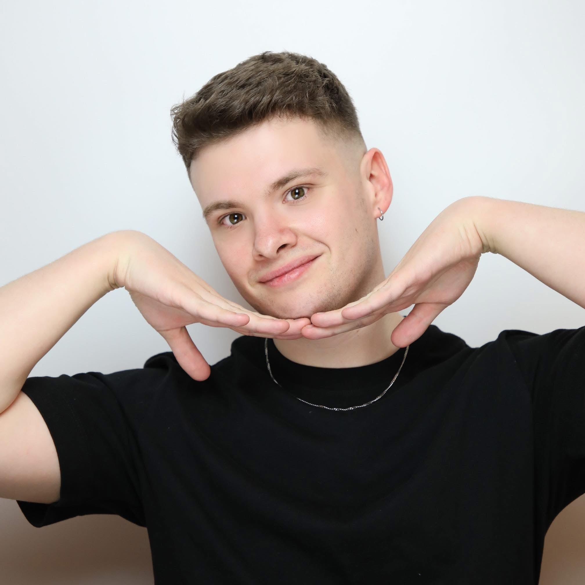 A man with brown hair wearing a black t-shirt. He has his hands in a pose under his chin.