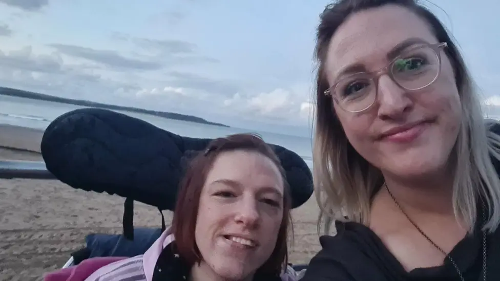 Sara is taking a picture of Vicky and herself. Vicky is in her wheelchair and looking at the camera. Sarah is standing next to her.