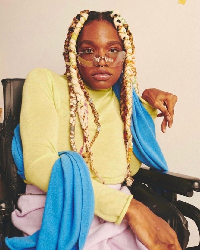 Aaron is posing, looking at the camera. She has blond and brown braids. She is wearing tinted glasses.