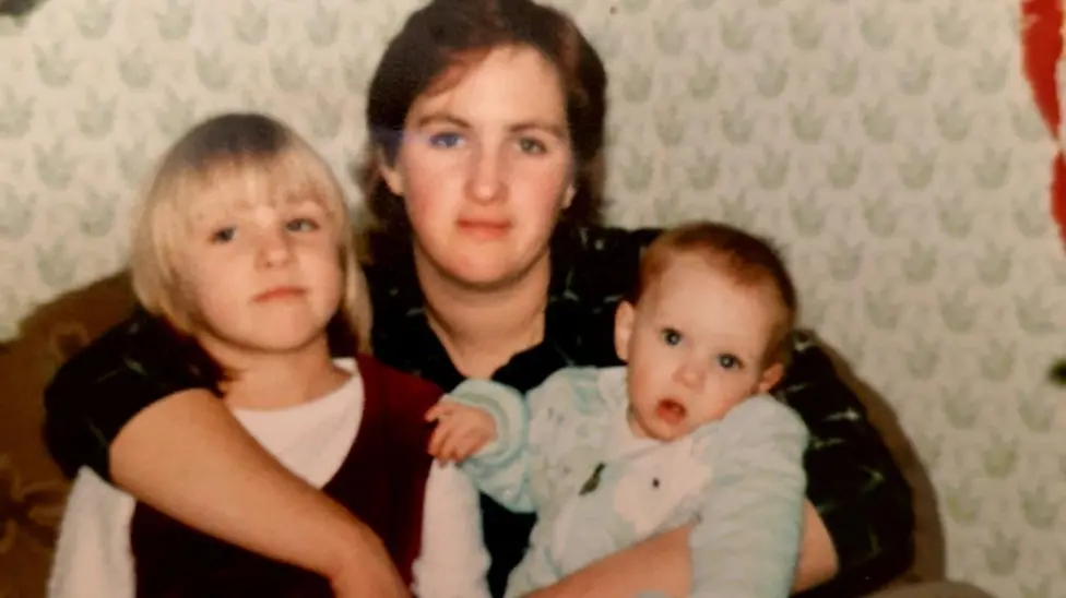 An old image of Sara, Vicky and their mum. Their mum is holding baby Vicky and toddler Sara. They are looking at the camera