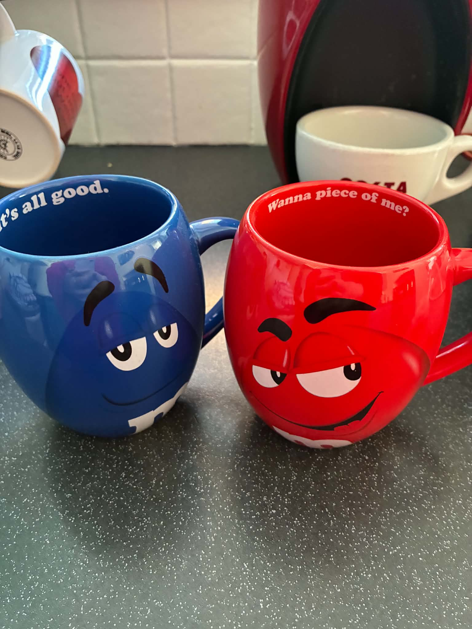 Two M&M's-branded ceramic coffee mugs sitting side-by-side on a dark countertop. The blue mug on the left features a cool, relaxed M&M character face and the text "It's all good." printed inside the inner rim. The red mug on the right features a smirking M&M character face with the text "Wanna piece of me?" printed along its inner rim. In the background, a white coffee cup sits under a red coffee machine against a white tiled backsplash.