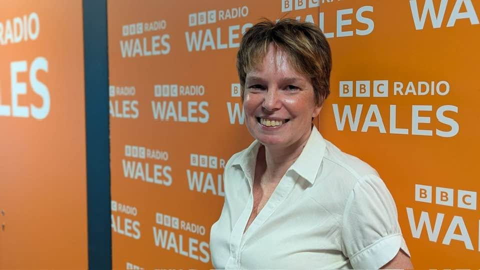 Sara is stood in front of BBC Wales logo sign. Sara has short brown hair and is wearing a white shirt.