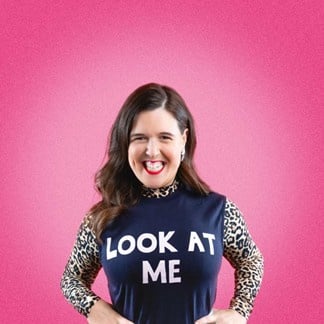 Rosie stands facing the camera. She is wearing a navy top with " look at me" in bold white letters.She has a long sleeve leopard print top underneath. Rosie has long dark hair. she is wearing red lipstick.