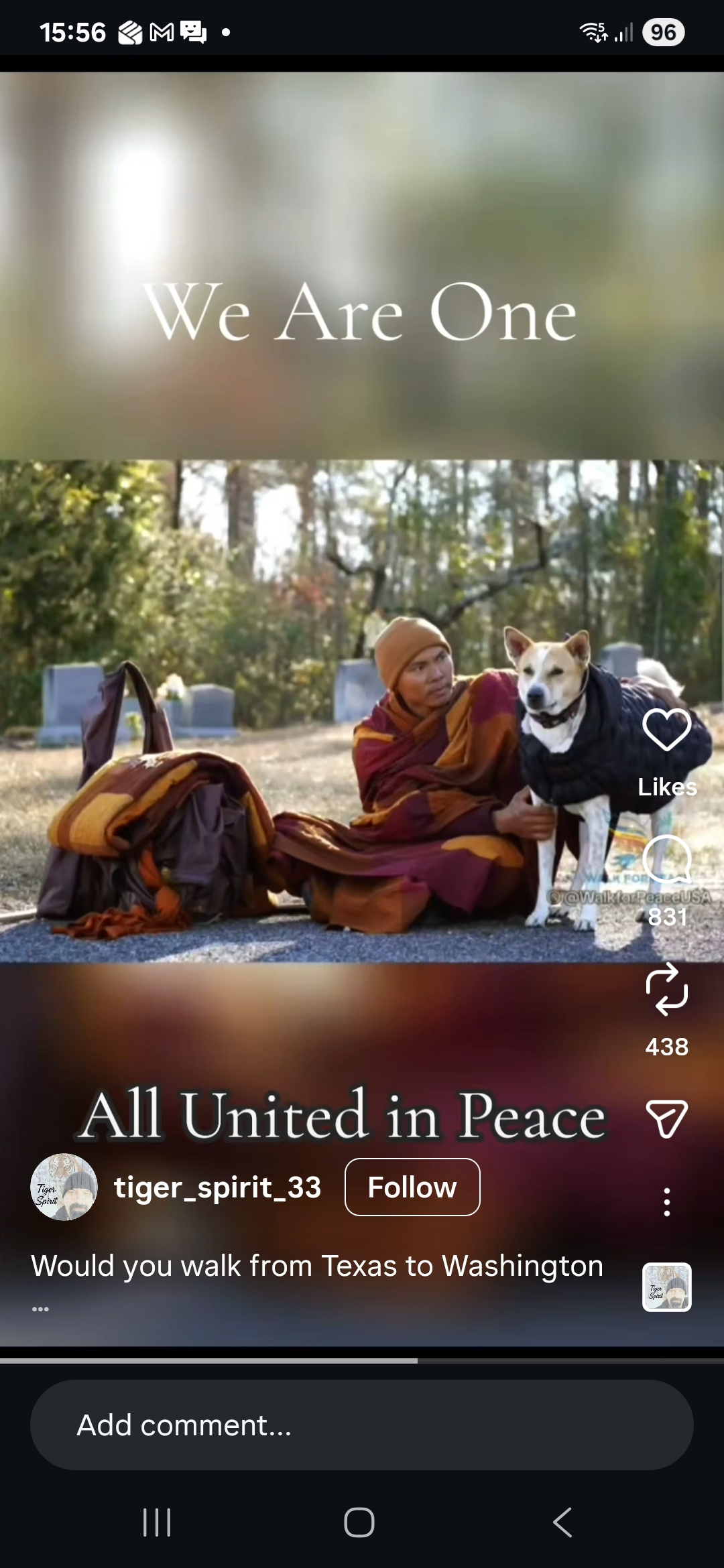 This is a screenshot from a phone showing a social media post. At the center of the image is a peaceful outdoor scene in what looks like a cemetery or memorial park. Trees with autumn leaves stand in the background, and gravestones are visible behind the subjects. In the foreground, a Buddhist monk is sitting on the ground. He is wearing layered robes in deep red, orange, and brown, along with a knit cap. Next to him is a medium-sized dog with tan and white fur. The dog is wearing a black jacket and standing calmly while the monk gently holds it close, as if comforting or protecting it. Beside them is a large bundled bag or backpack wrapped in fabric, suggesting they may be traveling. Over the image, there is white text that reads: “We Are One” and near the bottom: “All United in Peace” On the right side of the screen are social media icons showing likes, comments, and shares. The account name shown is “tiger_spirit_33,” and part of a caption reads: “Would you walk from Texas to Washington…” Overall, the image gives a feeling of calm, compassion, and unity between the monk and the dog in a quiet, reflective setting.
