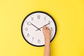 A black and white clock on a yellow background. Someone's hand is adjusting the time.