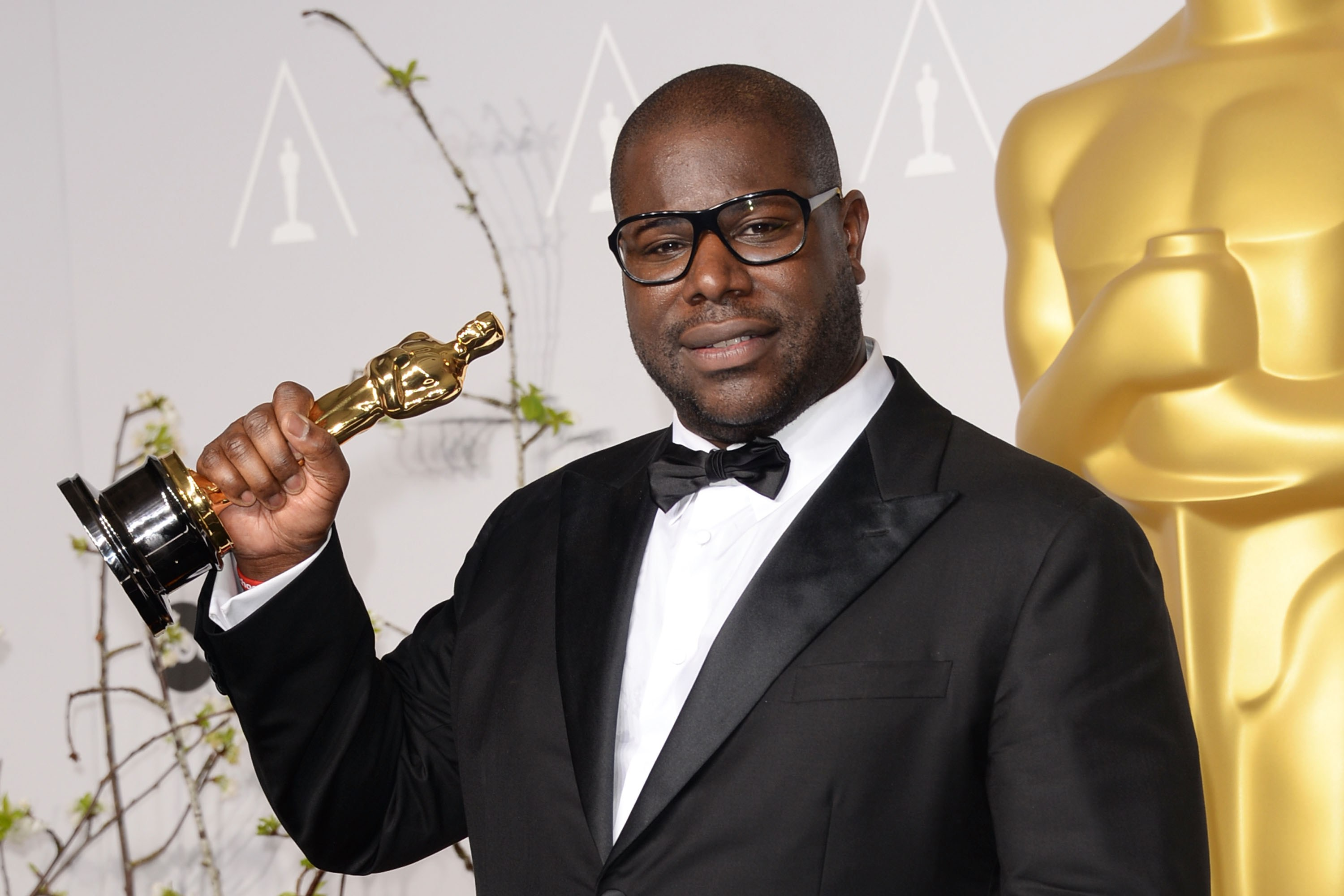 A photo of Sir Steve McQueen who is a black man, with black glasses wearing a very expensive and nice suit holding a BAFTA