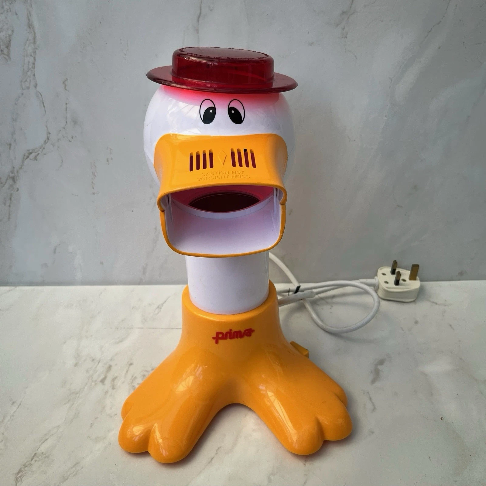 A prima popcorn maker in the shape of a duck. A white duck wearing a plastic red hat and with an orange beak and feet. The duck's bill is where the popcorn comes out.