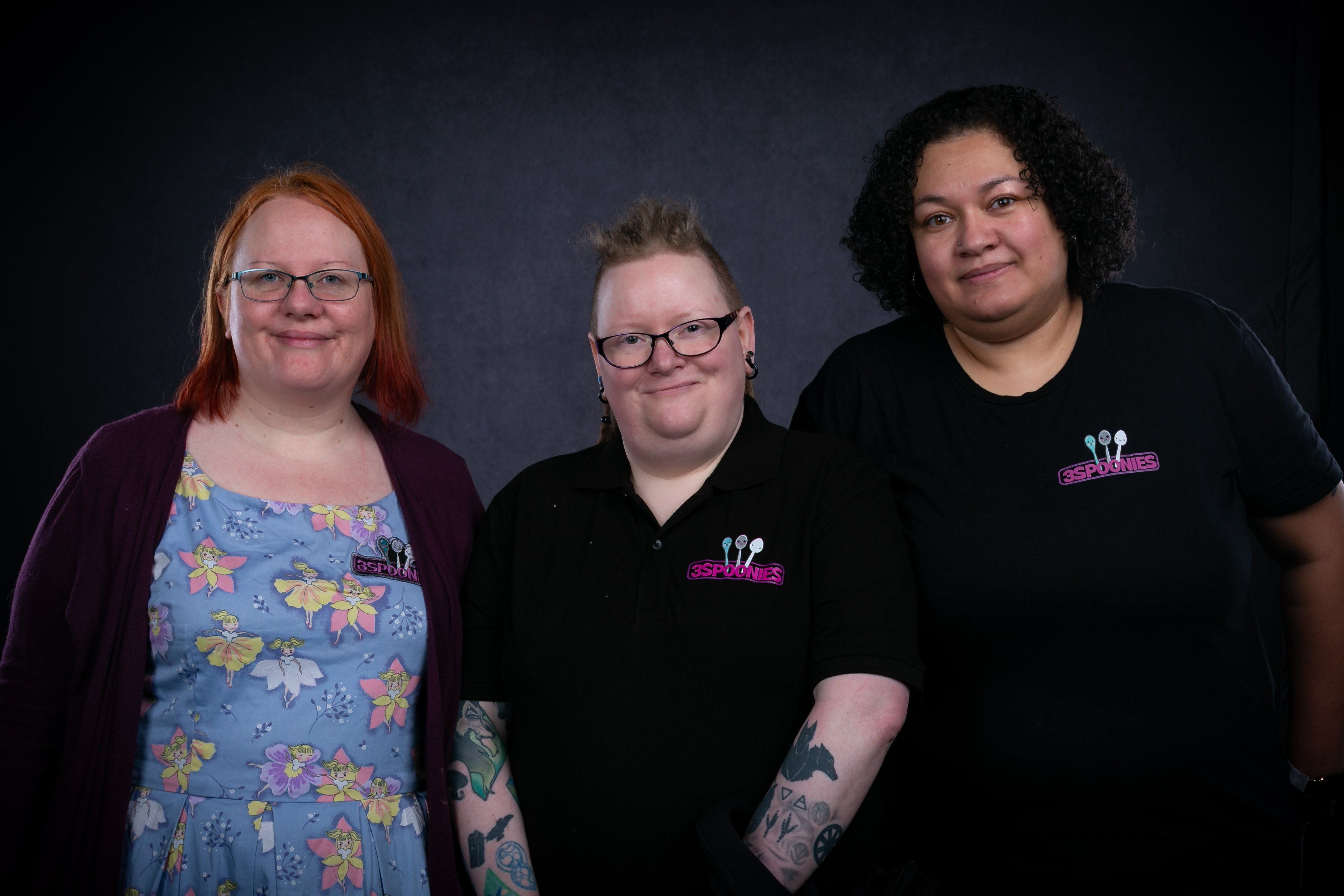 "Three people standing together against a dark background, smiling at the camera. Two wear black shirts with the purple “3Spoonies” logo, and one wears a light blue dress with a fairy pattern and a purple cardigan."