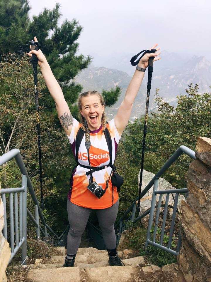 full body image of a young adult female with her arms in the air and smiling holding walking poles and wearing an orange and white MS society fundraising vest with mountain scenery in the background