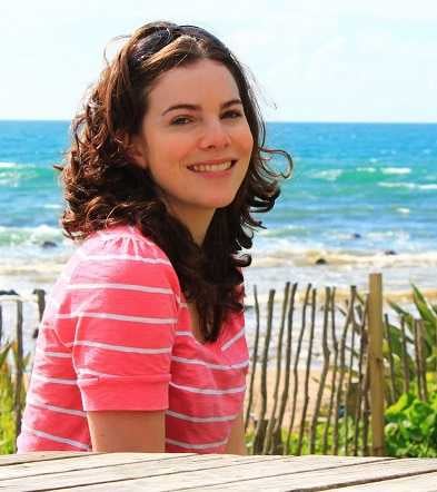 headshot of a woman with brown curly hair and wearing a pink top smiling at the camera with beach and ocean in the background