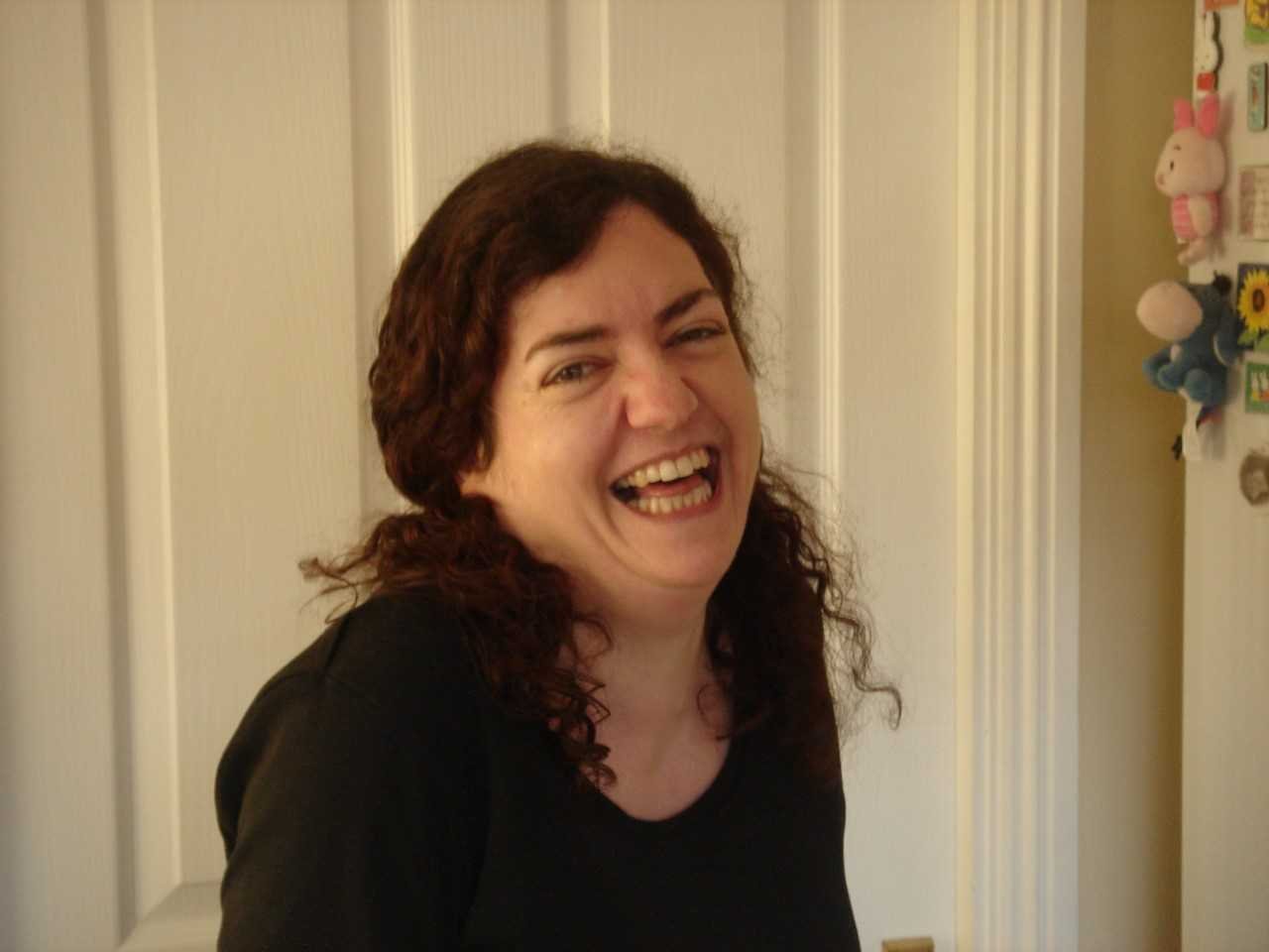 headshot of ruth with brown curly hair tied back and in a black top laughing and smiling at camera