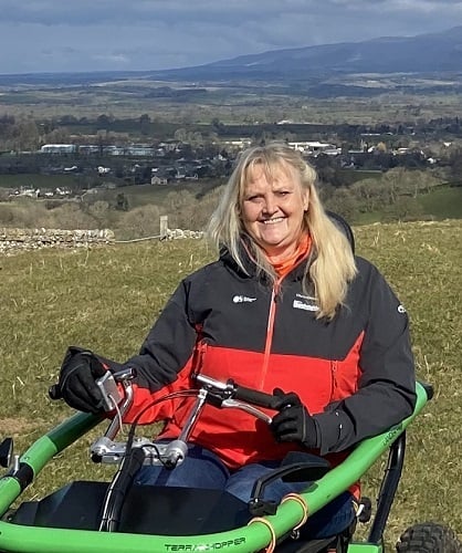 Debbie smiling sat in her wheelchair with green hills and countryside in the background