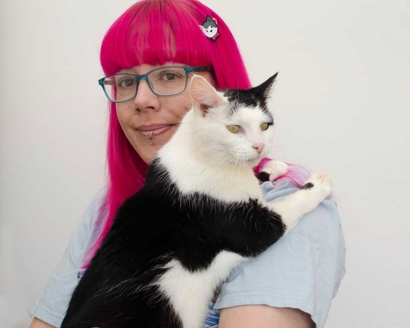 image of a girl with bright pink hair and wearing glasses holding a black and white cat over her shoulder